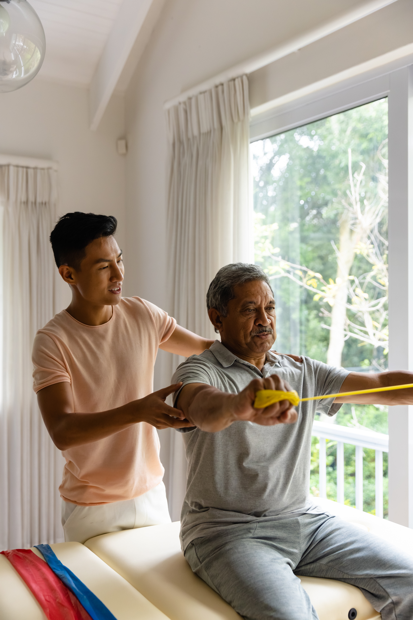 Happy diverse male physiotherapist advising and senior male patient using exercise band. Physiotherapy, healthcare, wellbeing, and senior lifestyle, unaltered.